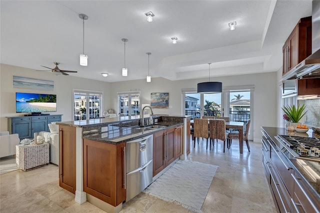 kitchen with sink, dark stone countertops, hanging light fixtures, a kitchen island with sink, and wall chimney range hood