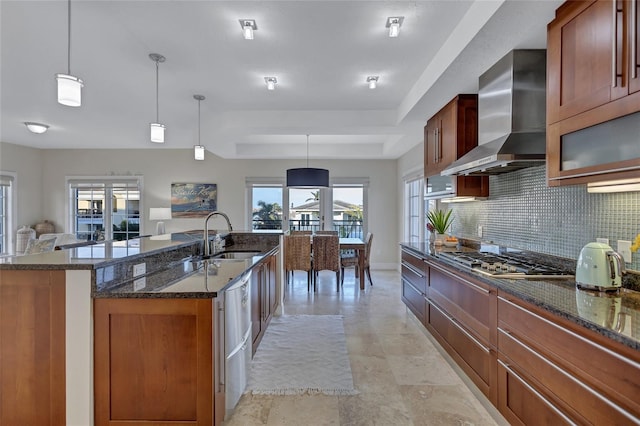 kitchen with wall chimney range hood, sink, a tray ceiling, an island with sink, and decorative light fixtures