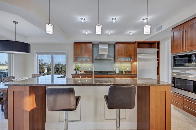 kitchen with a kitchen breakfast bar, dark stone counters, hanging light fixtures, built in appliances, and wall chimney exhaust hood