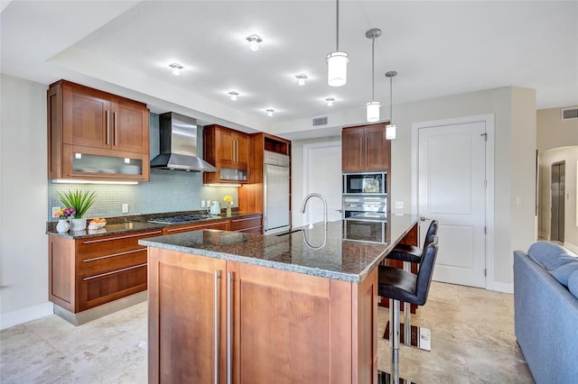 kitchen featuring wall chimney range hood, built in appliances, an island with sink, a kitchen bar, and decorative light fixtures