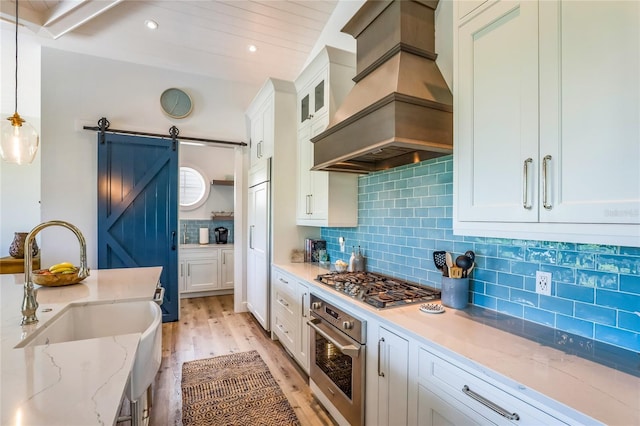 kitchen featuring appliances with stainless steel finishes, custom range hood, white cabinets, decorative light fixtures, and a barn door