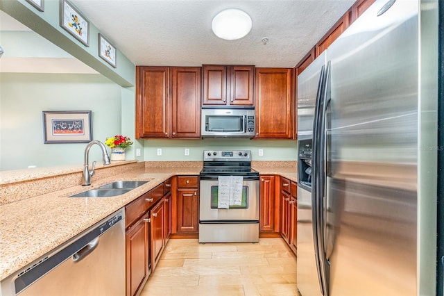 kitchen with a sink, a textured ceiling, brown cabinetry, light stone countertops, and stainless steel appliances
