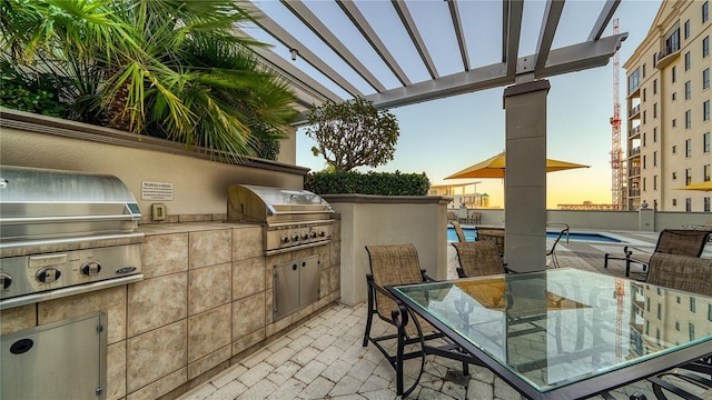 patio terrace at dusk featuring area for grilling, a community pool, and an outdoor kitchen