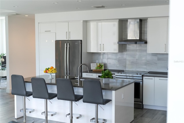 kitchen featuring wall chimney exhaust hood, white cabinetry, a center island with sink, stainless steel appliances, and backsplash