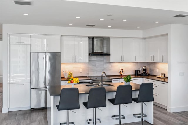 kitchen with wall chimney exhaust hood, sink, a center island with sink, stainless steel refrigerator, and white cabinets