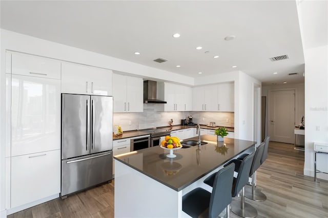 kitchen with white cabinetry, a center island with sink, appliances with stainless steel finishes, decorative backsplash, and wall chimney range hood