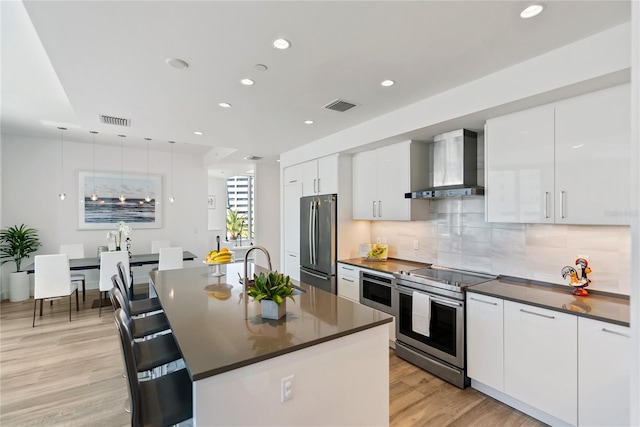 kitchen featuring wall chimney range hood, light wood-type flooring, stainless steel appliances, a kitchen island with sink, and white cabinets