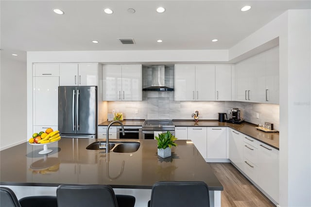 kitchen featuring wall chimney range hood, sink, stainless steel appliances, white cabinets, and a center island with sink