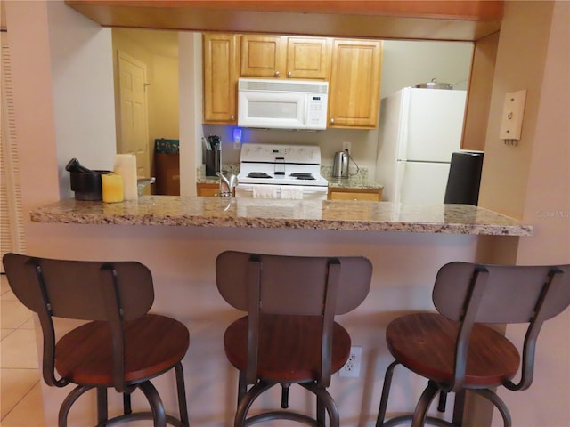 kitchen featuring light tile patterned flooring, a kitchen breakfast bar, light stone counters, light brown cabinets, and white appliances