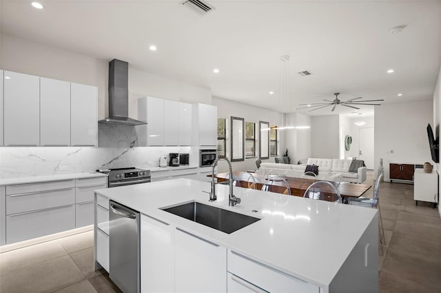 kitchen with an island with sink, white cabinetry, sink, stainless steel appliances, and wall chimney range hood