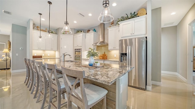 kitchen featuring wall chimney range hood, a center island with sink, white cabinets, and stainless steel refrigerator with ice dispenser