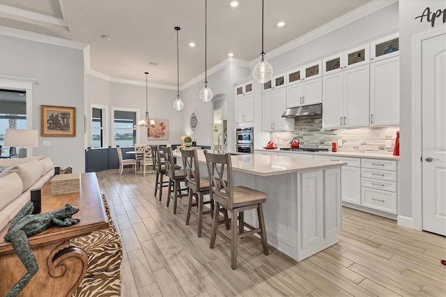 kitchen featuring decorative light fixtures, tasteful backsplash, white cabinetry, a kitchen island with sink, and light wood-type flooring