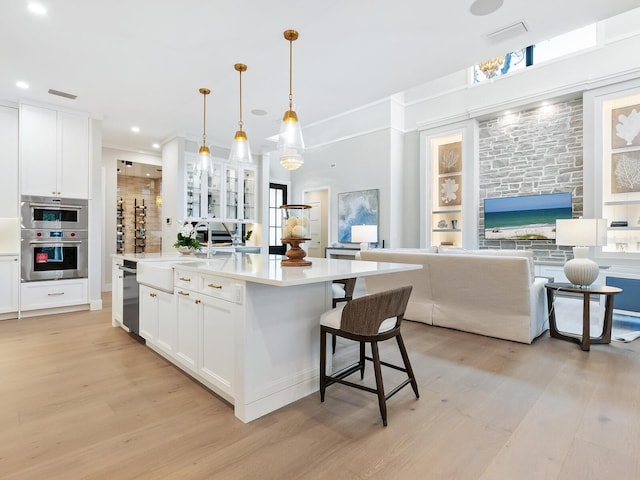 kitchen featuring an island with sink, white cabinets, decorative light fixtures, stainless steel double oven, and light wood-type flooring