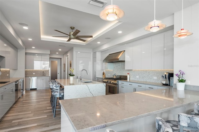 kitchen with high end stove, sink, hanging light fixtures, a tray ceiling, and wall chimney range hood