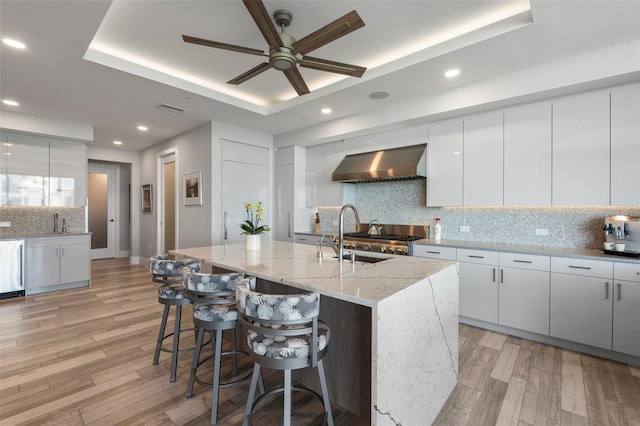 kitchen featuring sink, white cabinets, a kitchen island with sink, a tray ceiling, and wall chimney range hood