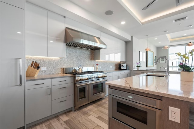 kitchen featuring pendant lighting, wall chimney range hood, sink, built in appliances, and light stone counters