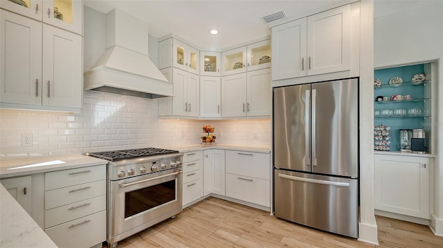 kitchen featuring appliances with stainless steel finishes, white cabinetry, custom exhaust hood, light stone countertops, and light hardwood / wood-style flooring