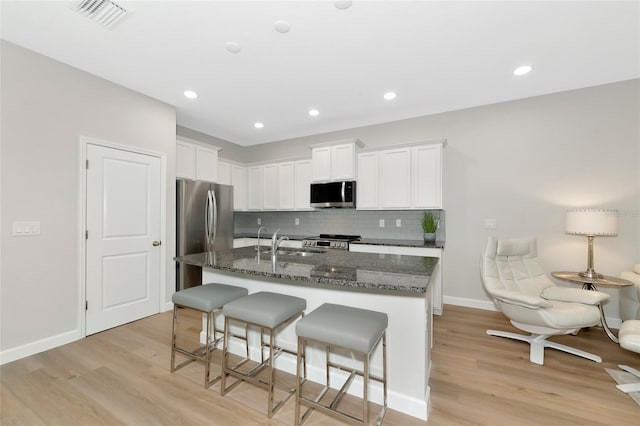 kitchen featuring white cabinetry, stainless steel appliances, dark stone countertops, and a center island with sink