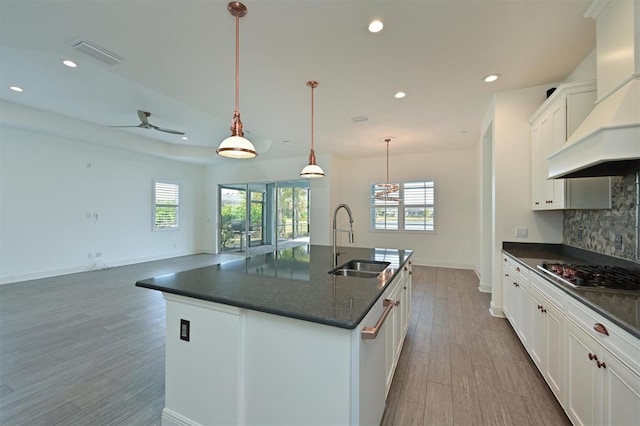 kitchen featuring sink, premium range hood, a kitchen island with sink, hanging light fixtures, and stainless steel gas cooktop