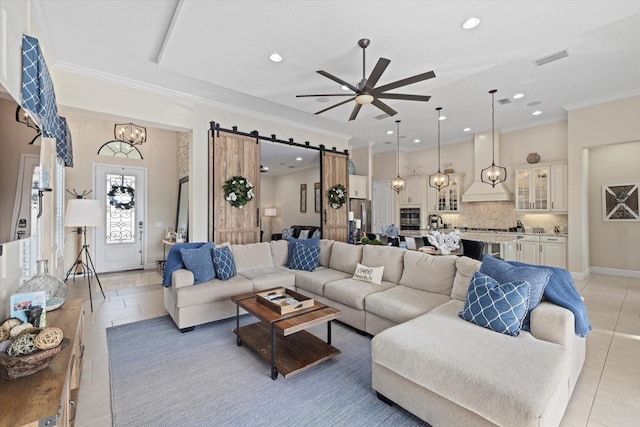 living room with crown molding, a barn door, ceiling fan with notable chandelier, and light tile patterned floors