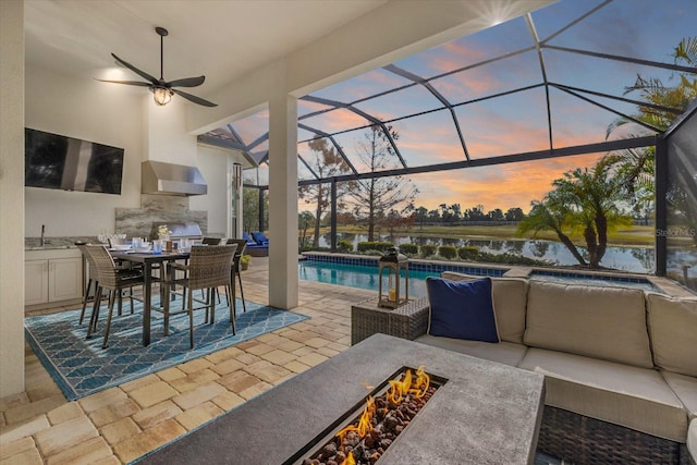 patio terrace at dusk featuring a wet bar, a water view, area for grilling, glass enclosure, and an outdoor living space with a fire pit