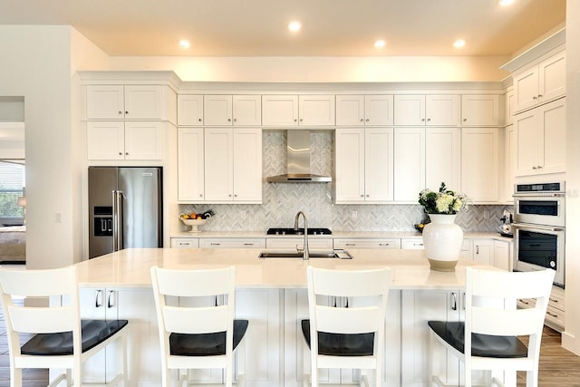 kitchen with wall chimney range hood, appliances with stainless steel finishes, a center island with sink, light stone countertops, and a kitchen bar