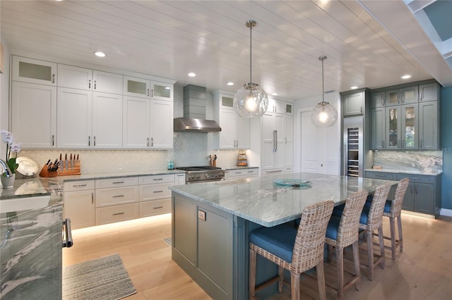 kitchen featuring a large island, wall chimney range hood, hanging light fixtures, light stone counters, and light hardwood / wood-style floors