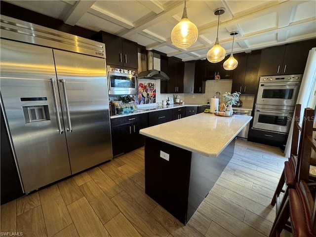 kitchen with decorative light fixtures, coffered ceiling, a center island, built in appliances, and wall chimney exhaust hood