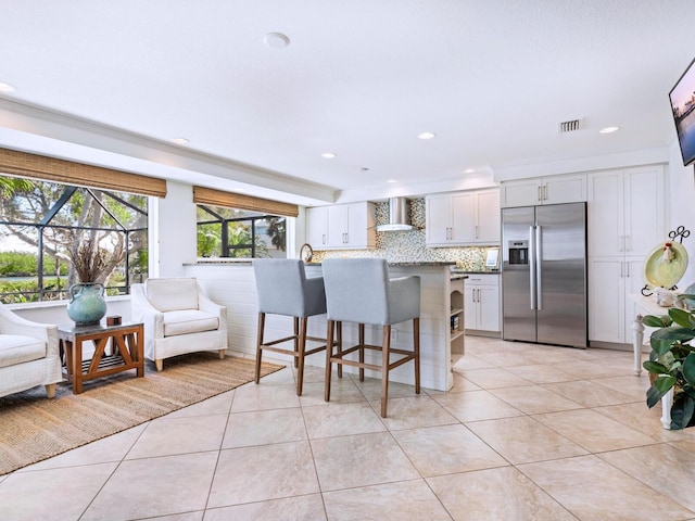 kitchen with wall chimney exhaust hood, a breakfast bar area, built in refrigerator, white cabinets, and backsplash