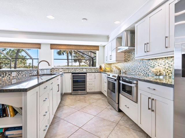 kitchen featuring wall chimney range hood, sink, dark stone countertops, white cabinets, and beverage cooler