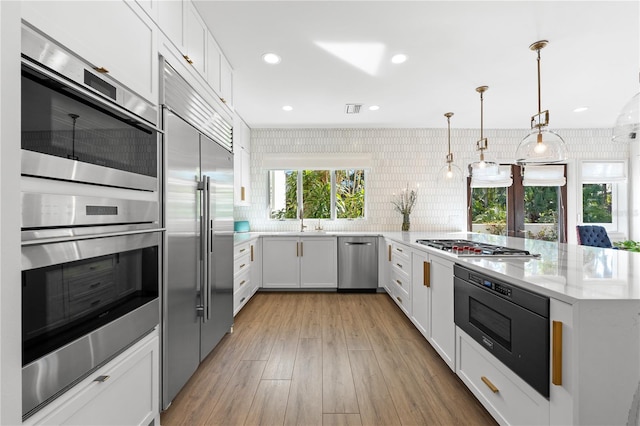 kitchen featuring tasteful backsplash, white cabinetry, hanging light fixtures, built in appliances, and light hardwood / wood-style flooring