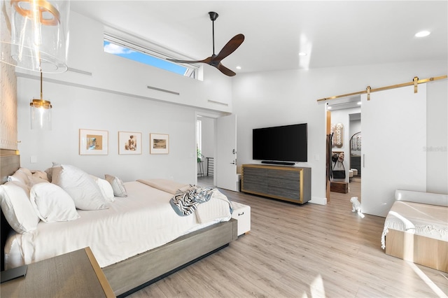 bedroom with ceiling fan, a barn door, light hardwood / wood-style flooring, and a high ceiling