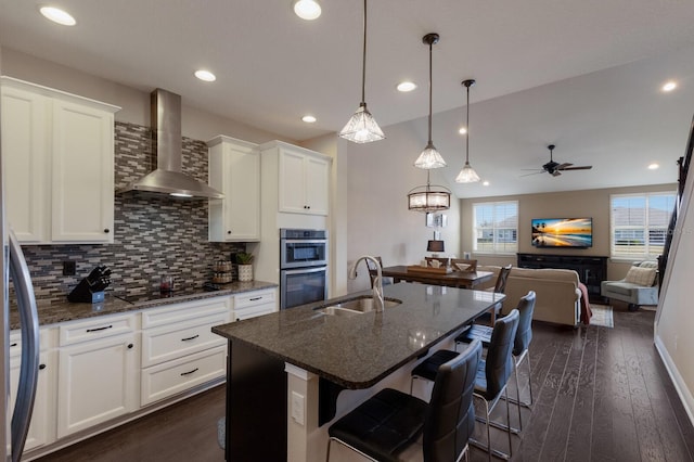 kitchen featuring wall chimney exhaust hood, sink, white cabinetry, dark stone counters, and a kitchen island with sink