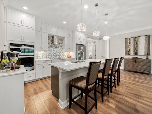 kitchen with pendant lighting, sink, white cabinets, a kitchen island with sink, and wall chimney range hood