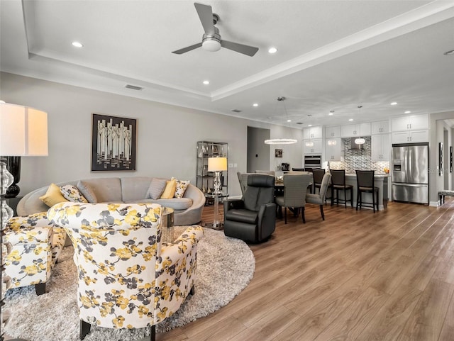 living room with a raised ceiling, ceiling fan, and light wood-type flooring