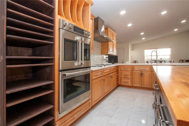 kitchen featuring sink, wooden counters, stainless steel double oven, wall chimney range hood, and backsplash