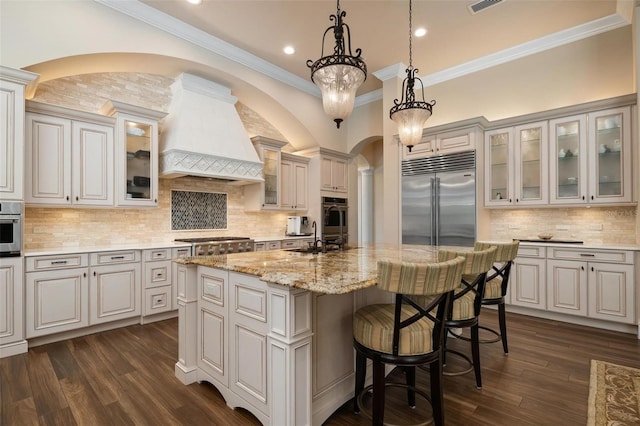 kitchen with premium range hood, hanging light fixtures, an island with sink, stainless steel appliances, and light stone countertops