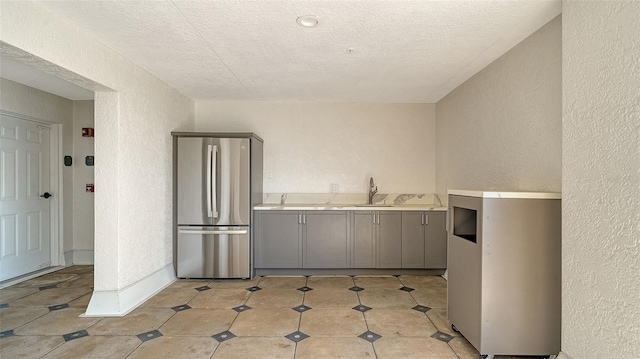 kitchen featuring light countertops, a textured wall, gray cabinetry, freestanding refrigerator, and a sink