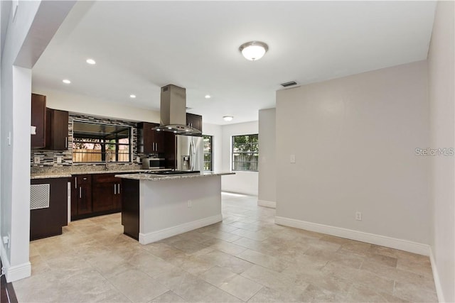 kitchen with stainless steel refrigerator with ice dispenser, island range hood, light stone countertops, a kitchen island, and decorative backsplash