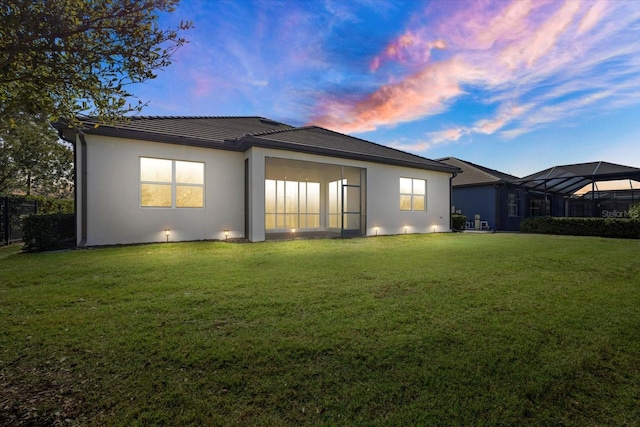 back house at dusk featuring glass enclosure and a lawn