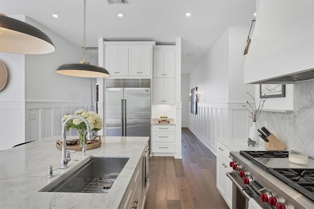 kitchen featuring a wainscoted wall, high end appliances, white cabinets, a sink, and light stone countertops