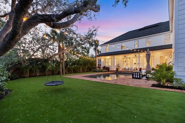 back of house at dusk with a ceiling fan, an outdoor pool, a lawn, and a patio