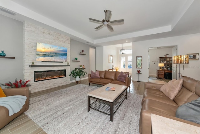 living room with hardwood / wood-style flooring, a stone fireplace, a raised ceiling, and ceiling fan