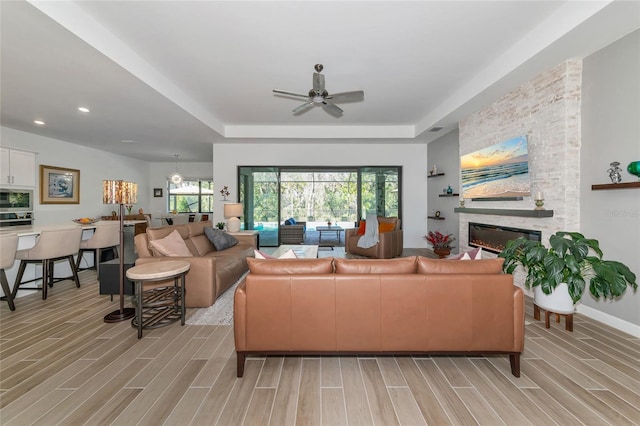 living room with a stone fireplace, a raised ceiling, and ceiling fan with notable chandelier