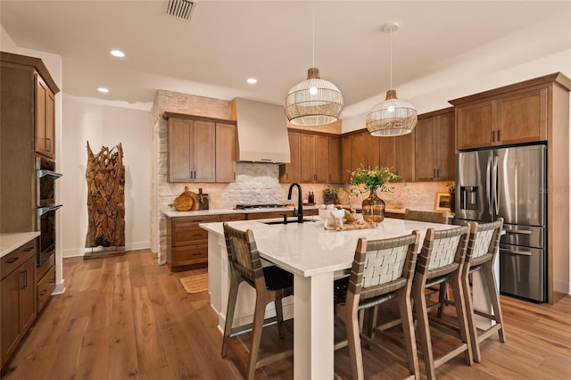 kitchen with a center island with sink, decorative light fixtures, stainless steel appliances, premium range hood, and a sink