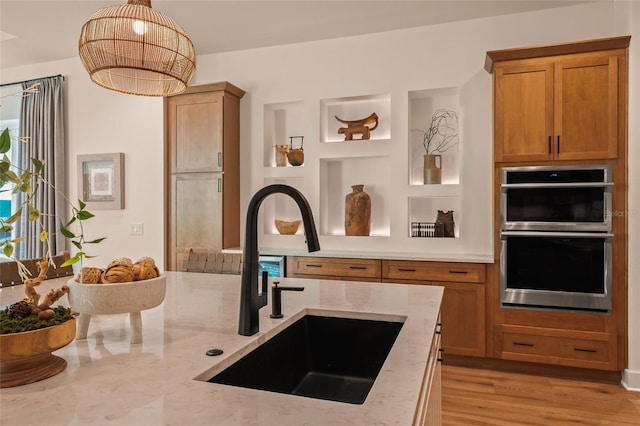 kitchen with light stone counters, stainless steel double oven, a sink, light wood-type flooring, and decorative light fixtures