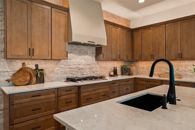 kitchen with light stone countertops, stainless steel gas cooktop, premium range hood, a sink, and decorative backsplash