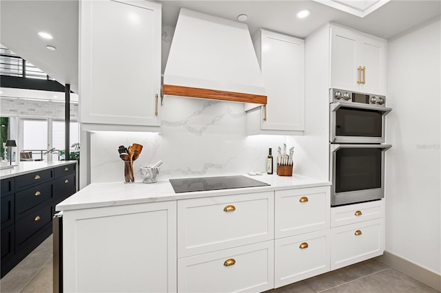 kitchen featuring decorative backsplash, double oven, white cabinetry, premium range hood, and black electric cooktop