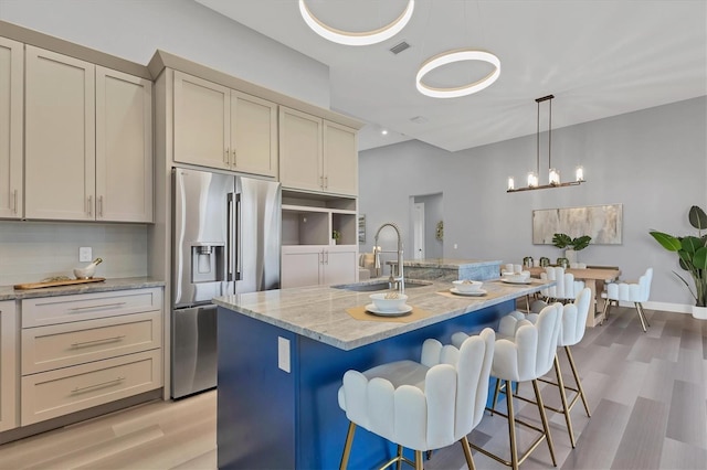 kitchen with sink, a kitchen island with sink, stainless steel refrigerator with ice dispenser, light stone counters, and cream cabinetry