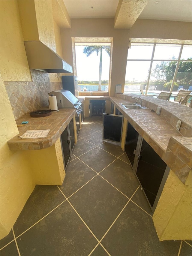 kitchen with wall chimney range hood, tile counters, dark tile patterned floors, a textured wall, and a sink